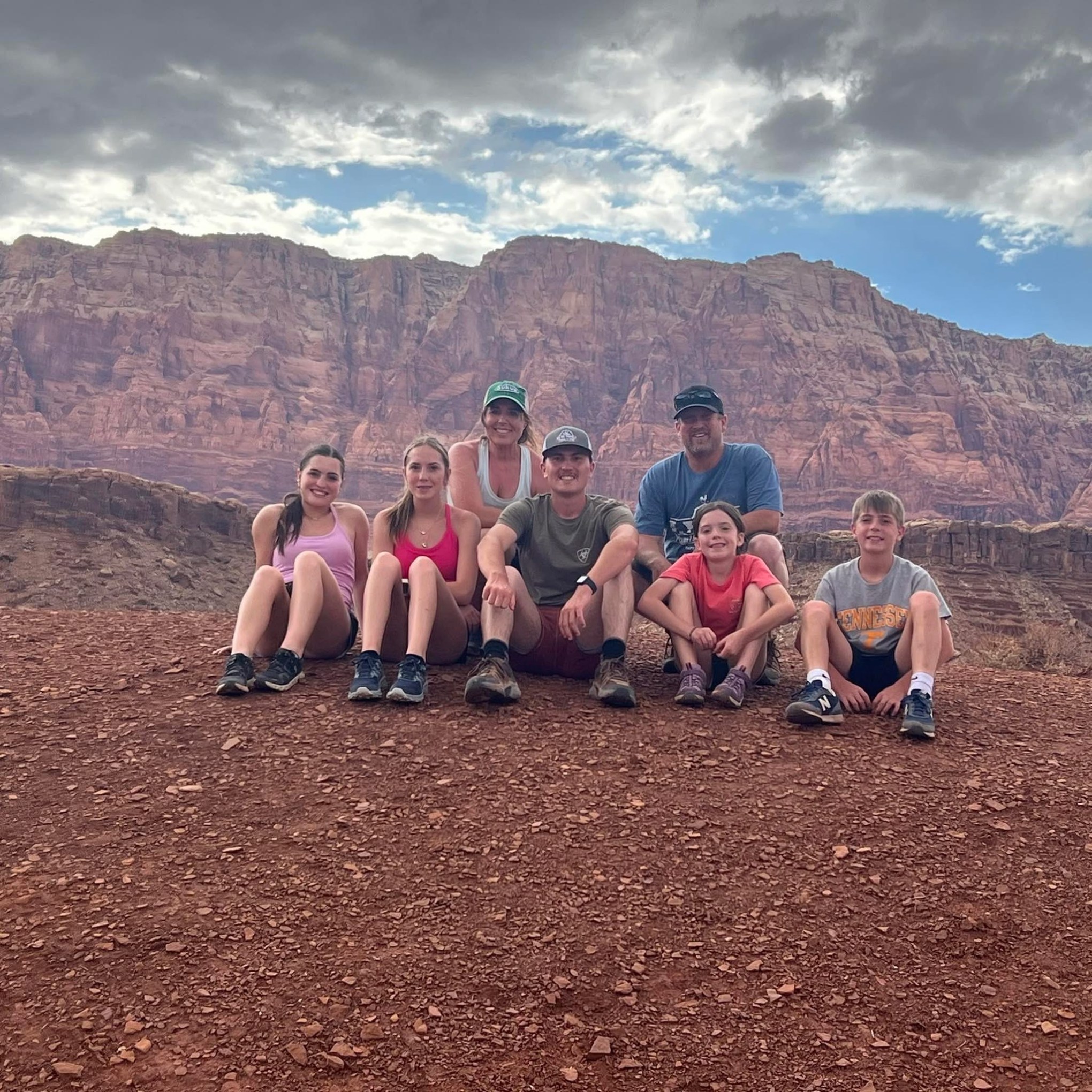A group of seven people sitting on a rocky terrain with red cliffs in the background.