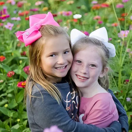 Two smiling girls with bows hugging in a colorful flower garden.