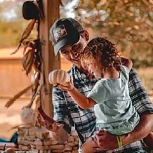 Man holding a child, both looking at a small pumpkin in a rustic outdoor setting.