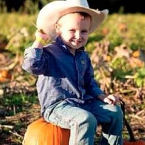 Smiling child in cowboy hat sitting on pumpkin in a field.